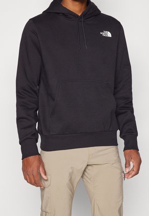 Mann trägt einen schwarzen The North Face Hoodie mit Fronttasche und beigen Cargohosen, gezeigt vom Hals bis zur Hüfte.