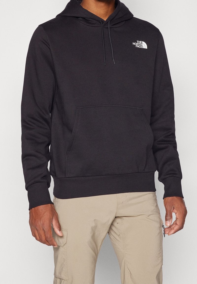 Homme portant un sweat à capuche noir The North Face avec une poche avant et un pantalon cargo beige, montré du cou aux hanches.