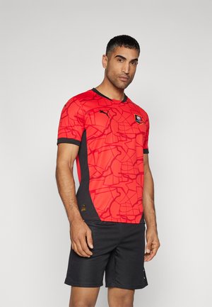 Maillot rouge à manches courtes avec un motif géométrique, accents noirs et col rond. Porté avec un short noir. Logo Puma sur la manche.