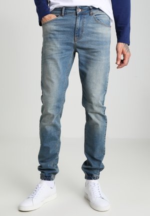 Homme portant un jean jogger en denim bleu délavé avec des poignets élastiques, des baskets blanches et une chemise à manches longues bleu marine et blanche, debout la main dans la poche.