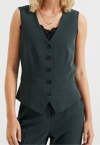 Gilet sans manches vert foncé avec un col en V, six boutons noirs, deux poches latérales et une coupe ajustée, fabriqué en tissu lisse.
