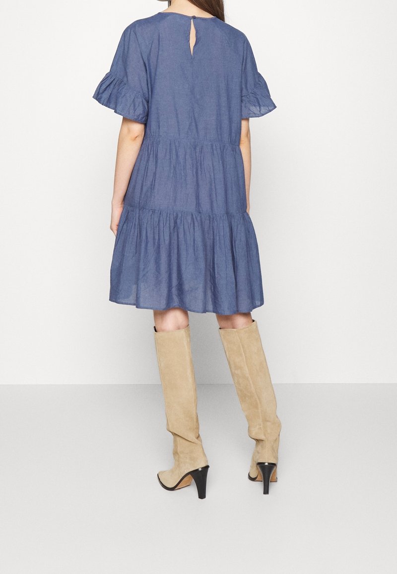 Femme portant une robe bleue longueur genou avec des manches courtes à volants et des bottes beige montantes à talons hauts, montrée de dos sur un fond uni.
