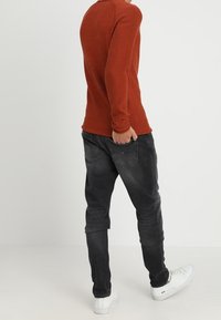 Czerwona koszula z długim rękawem i zaokrąglonym dekoltem, zestawiona z ciemnoszarymi zwężanymi jeansami i białymi trampkami, widziana od tyłu.