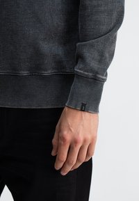 Donkergrijze sweater met geribde manchetten en zoom, met een textuurafwerking. Kleine labeldetail op de manchette. Hand rustend op zwarte broek.