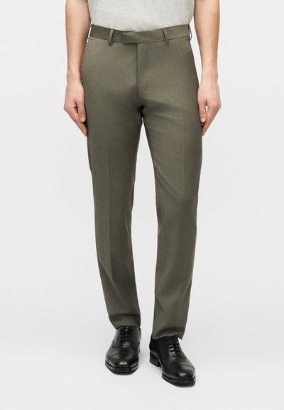 Άνδρας που φοράει παντελόνι φόρμας ελιάς με πτυχή, μαύρα δερμάτινα παπούτσια και ανοιχτό γκρι πουκάμισο σε λευκό φόντο.