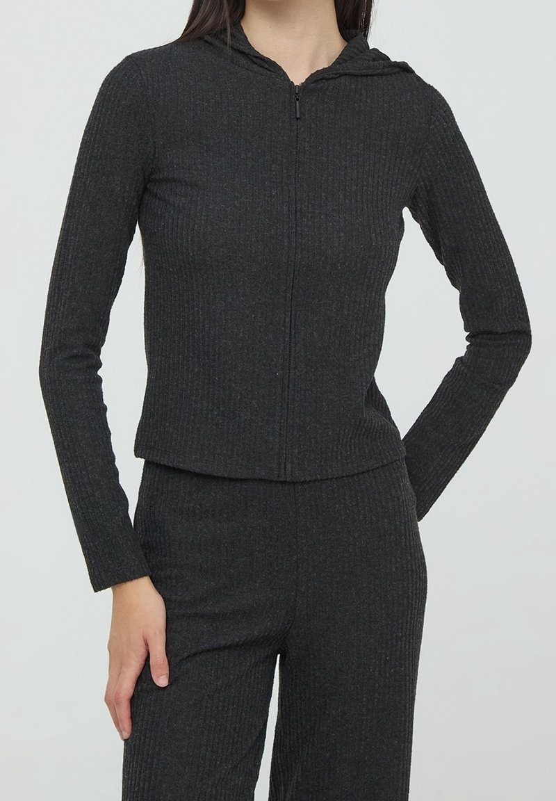 Femme portant un sweat à capuche zippé côtelé gris foncé et un pantalon côtelé assorti, la main gauche posée sur la hanche, sur un fond uni.