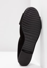 Chaussure en suede noire avec un talon légèrement surélevé et une semelle en caoutchouc texturé, présentant des rainures pour la traction. Accents comprenant une fermeture sécurisée.