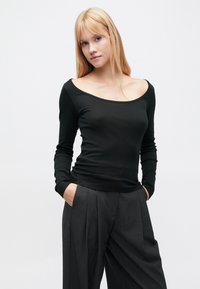 Μακρυμάνικη μαύρη μπλούζα με φαρδιά λαιμόκοψη σε σχήμα scoop, εφαρμοστό μπούστο και απαλή υφή, συνδυασμένη με ψηλόμεσα γκρι παντελόνια με πλισέ.