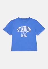 Modré bavlněné tričko s krátkými rukávy. Na přední straně má kulatý nápis "STADIUM // PLAY" v růžové barvě a "1986" v sportovním designu. Texturovaná látka.