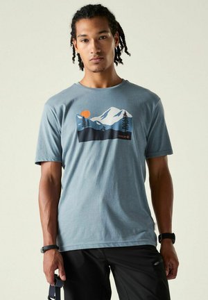 Mann mit geflochtenem Haar, der ein blaues T-Shirt mit einer Berg- und Baumgrafik, schwarze Hose und eine Uhr trägt, vor einem einfarbigen Hintergrund steht.