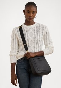 Schwarze Leder-Umhängetasche mit geprägt Logo, flaches Design, verstellbarem Gurt und goldfarbenen Hardware-Details. Trägt man über einem weißen Strickpullover.