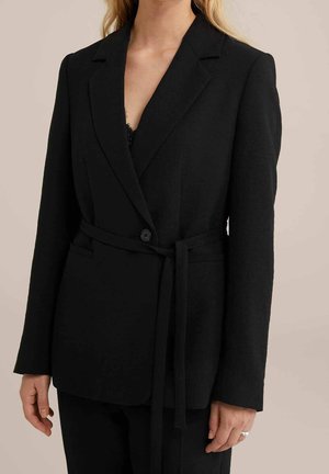 Vrouw draagt een zwarte, op maat gemaakte blazer met een enkele knoop en stoffen riem, gecombineerd met een zwarte broek tegen een neutrale achtergrond.