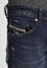 Jeans de mezclilla desgastados de color azul oscuro con corte slim, que presentan un bolsillo trasero visible con una etiqueta de la marca y detalles de remaches de cobre.