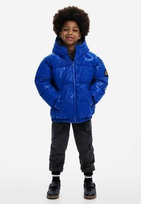 Blå dunjakke med blank finish, lynlås foran og hætte; kombineret med mørke bukser med mønster og sorte sko. Let polstret for varme.