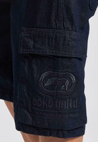 Tumman farkkukankaan cargo-shortsit sisältävät sävyyn painetun logo-patchin, etutaskut ja rennon istuvuuden, jossa on näkyviä sauma-akkentteja.