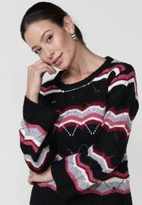 Pull noir avec un motif en zigzag rouge, gris et blanc ; présente une coupe ample, une texture tricotée et des manches larges avec un col rond.