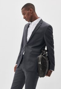 Traje gris carbón con un diseño estructurado, llevado sobre una camisa blanca. Se sostiene bajo el brazo un bolso de cuero negro con cierre de cremallera.