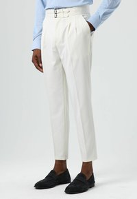 White pleated trousers with a high waistband featuring double metal buckles. Worn with black suede loafers, complemented by a light blue shirt.