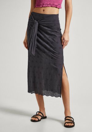 Kvinde iført sort midinederdel med blomstret hulmønster, sidelange slidser, bundet knude ved taljen, kombineret med sorte sandaler med stropper og en pink crop top.