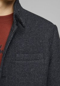 Texturerad mörkgrå jacka med ett subtilt rutmönster, med en liten bröstficka och en tryckknappskrage. Bärs över en röd skjorta.