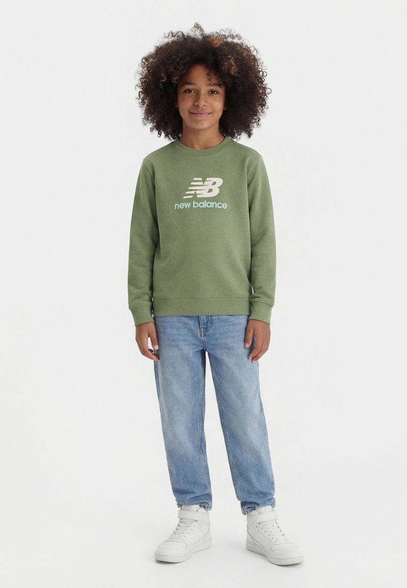 Παιδί με σγουρά μαλλιά που φοράει πράσινο φούτερ New Balance, ανοιχτόχρωμα μπλε τζιν και λευκά ψηλά αθλητικά παπούτσια, στέκεται μπροστά σε λευκό φόντο.