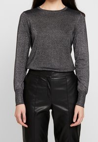 Långärmad svart topp i en glittrig, texturerad tyg. Har rund halsringning och en figurnära stil, kombinerad med svarta läderbyxor.