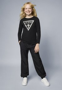 T-shirt noir à manches longues avec un logo triangulaire blanc portant "GUESS USA" au centre, associé à un pantalon noir ample et des baskets blanches.