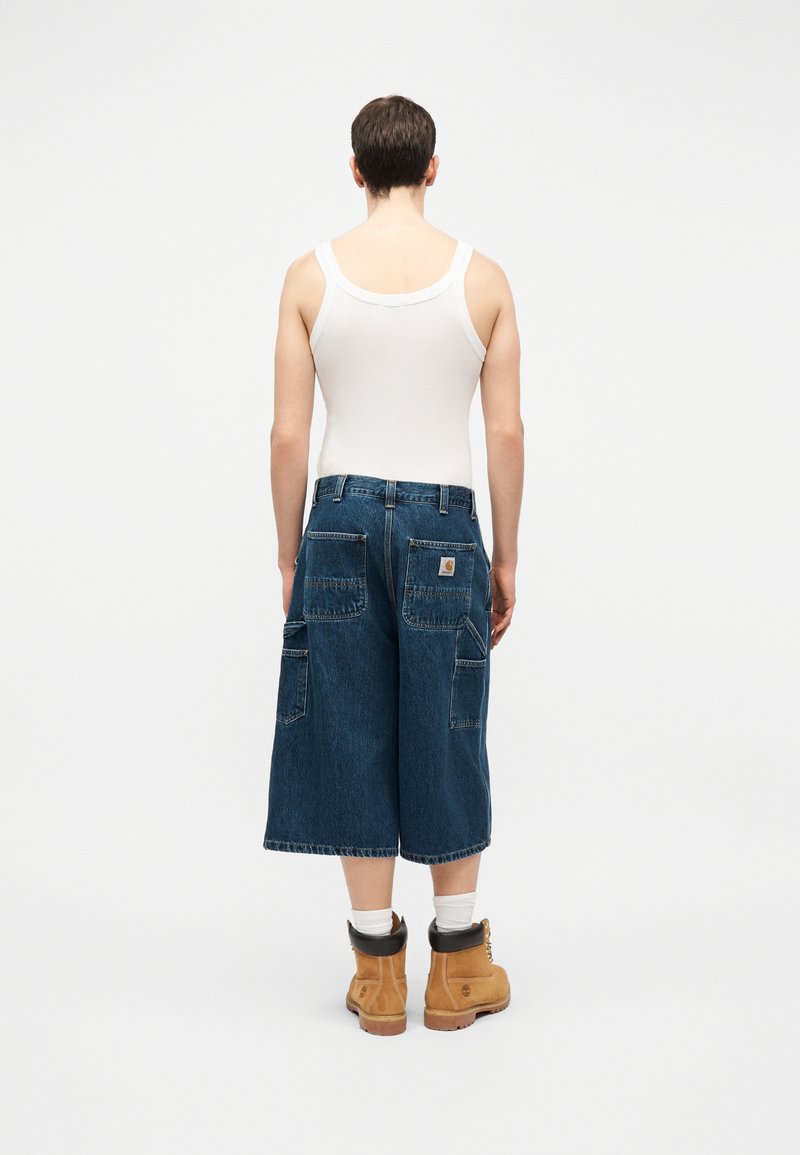Person står med ryggen til, iført hvid tanktop, blå vide cropped denimbukser, hvide sokker og brune arbejdstøvler på ensfarvet baggrund.