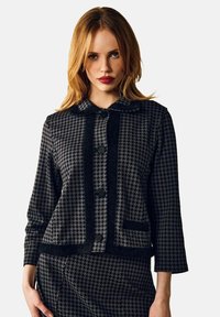 Sort og grå houndstooth-jakke med krave, tre knapper og en brystlomme. Struktureret stof med en tætsiddende pasform.