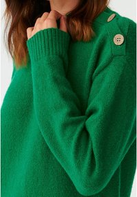 Pull vert en tissu texturé, avec des poignets et des épaules côtelés et des boutons en bois visibles. Détails en gros plan mis en avant.