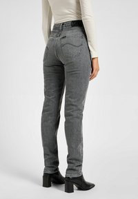 Jeans de denim gris con un ajuste slim, que presentan una cintura alta, bolsillos traseros y detalles de costura vertical, combinados con botas negras de tacón cuadrado.