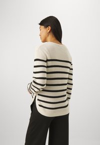 Kremgenser med svarte horisontale striper, ribbestruktur og lange ermer. Har en avrundet halslinje og sidespalter i kanten.