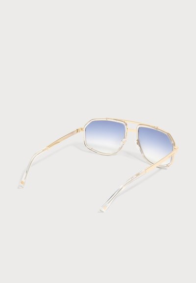 Lunettes de soleil à monture métallique dorée avec verres à dégradé bleu et extrémités de branches en plastique transparent sur un fond clair uni.