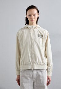Casaco com capuz em creme claro, com punhos e baixo elásticos. Apresenta um fecho de correr e um detalhe com logo no lado esquerdo. Material suave e leve.