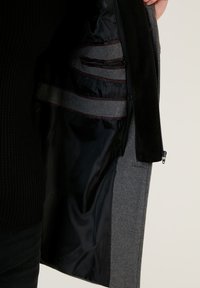 Tejido exterior de lana gris, forro de satén negro, con bolsillos con cremallera y bandas de tela gris en contraste. La textura se presenta suave y estructurada.