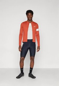 Férfi narancssárga kerékpáros dzsekiben, fehér pólóval, fekete kerékpáros nadrággal, fekete zokniban és fekete kerékpáros cipőben, sima háttér előtt áll.