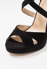 Chaussure à talon haut en daim noir avec bout ouvert, brides croisées et semelle compensée ; doublure intérieure lisse et détails de couture subtils.