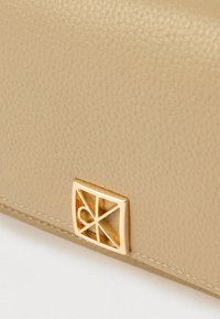 Επιφάνεια από μπεζ δέρμα με υφή και χρυσό γεωμετρικό λογότυπο στη κάτω γωνία. Ορατή απλή ραφή.