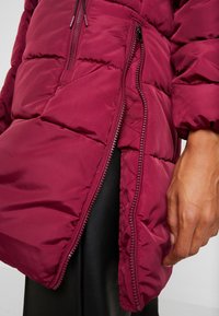 Burgunderfarbene Steppjacke mit gestepptem Design, ausgestattet mit einer Fronttasche mit Reißverschluss und einer glatten, leichten Textur. Detailaufnahme von Ärmel und Hand.