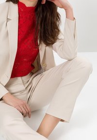 Beżowy, dopasowany garnitur z marynarką na miarę i skróconymi spodniami. Pod spodem czerwony koronkowy top dodaje faktury. Strój na jednolitym tle.