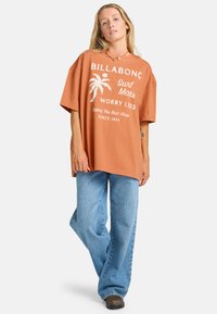 Camiseta de terracota de gran tamaño con gráfico de palmera blanca y texto, combinada con jeans de pierna ancha de color azul claro y zapatos negros.