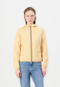 Giacca a cappuccio giallo chiaro con zip frontale, polsini elasticizzati e un piccolo logo. Indossata sopra jeans blu, texture liscia con dettagli minimi.