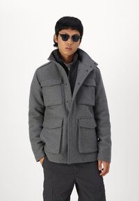 Chaqueta de mezcla de lana gris con alto cuello, cuatro bolsillos frontales y cierres de botones a presión. Llevada sobre un chaleco negro, combinada con pantalones oscuros.