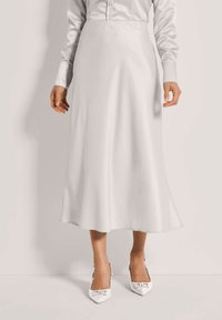 Woman wearing a long white satin skirt, white buttoned shirt, and white pointed-toe heels with buckle detail, standing against a plain background.