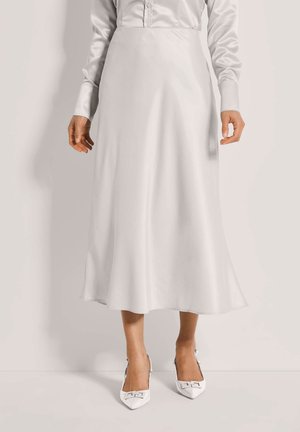 Frau trägt einen langen weißen Satinh Rock, ein weißes Hemd mit Knöpfen und weiße spitze High Heels mit Schnallendetail, stehend vor einem schlichten Hintergrund.