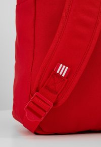 Red fabric backpack featuring a rectangular shape, adjustable strap with a plastic buckle, and white accent stripes. Textured surface.