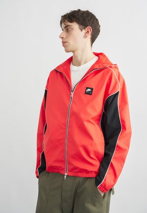 Ung mand iført en rød og sort Nike Air lynlåstrøje over en hvid skjorte, stående med hænderne i lommerne, kigger mod venstre mod en ensfarvet baggrund.