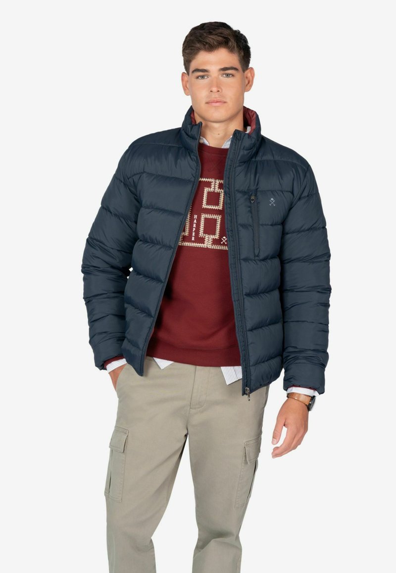 Chaqueta de plumas azul marino, diseño acolchado, cuello alto, bolsillos con cremallera, combinada con sudadera color burdeos y pantalones cargo beige.