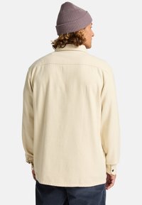Crème kleurig, textuur shirt met lange mouwen, voorzien van een knoopsluiting op de kraag en de manchetten. Draagt een lichtpaarse gebreide muts.