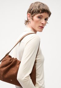 Braune Wildledertasche mit gerafftem Oberteil, Lederträger und goldenen Hardwaredetails. Das Modell trägt ein eng anliegendes cremefarbenes Hemd, das das Design der Tasche zur Geltung bringt.